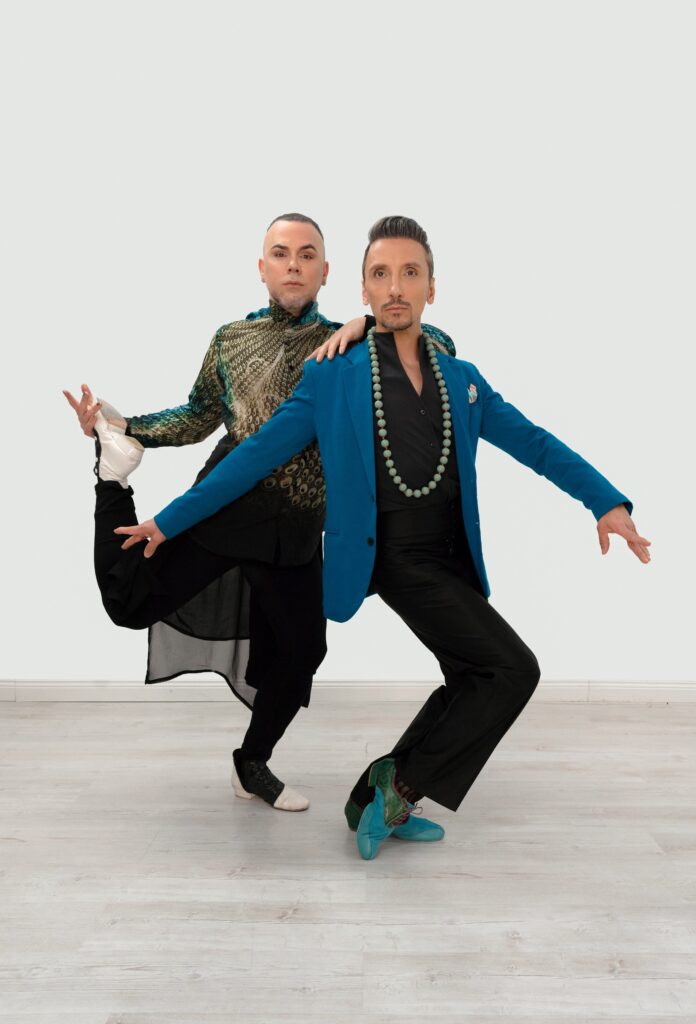 Two dancers in close hold, one taking the other’s weight, on a pale studio ground.